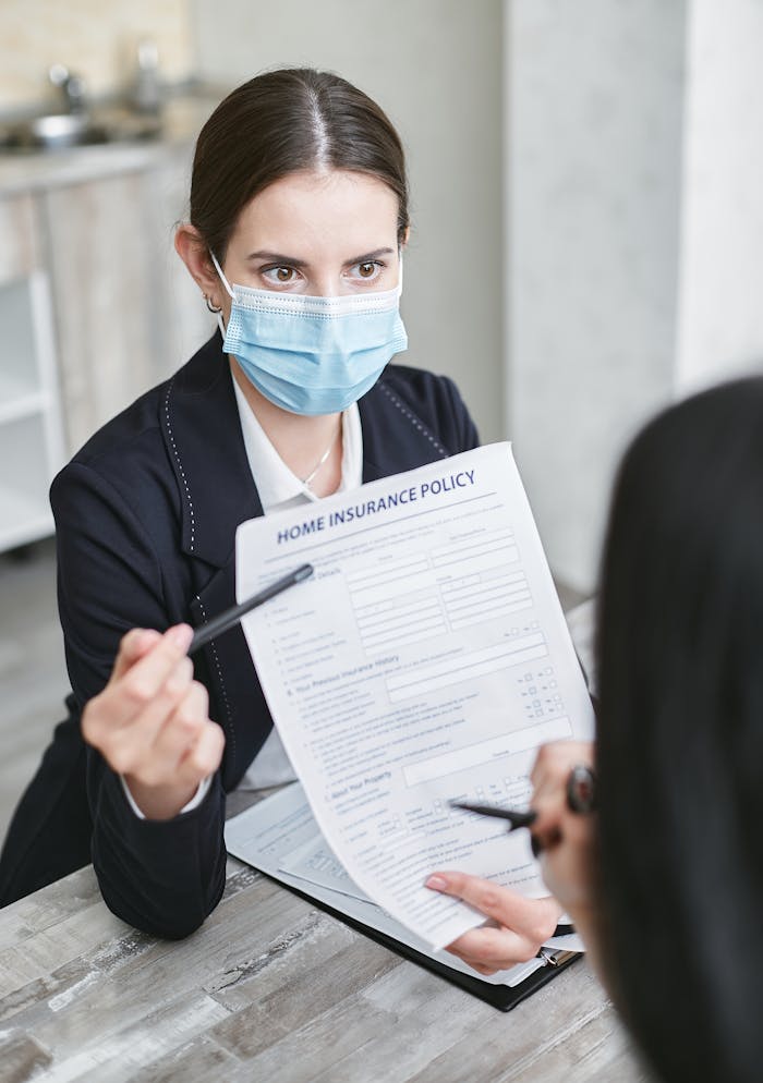 services-01 Professional woman wearing a face mask discussing a home insurance policy with a client at a desk.